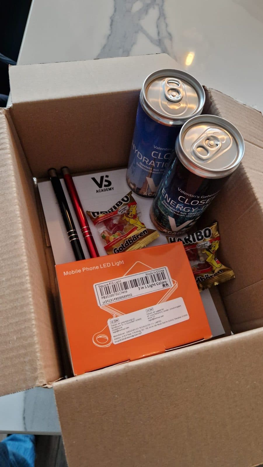 Blick in den Karton mit zwei Dosen, Stiften, Block, Haribo und LED-Licht
